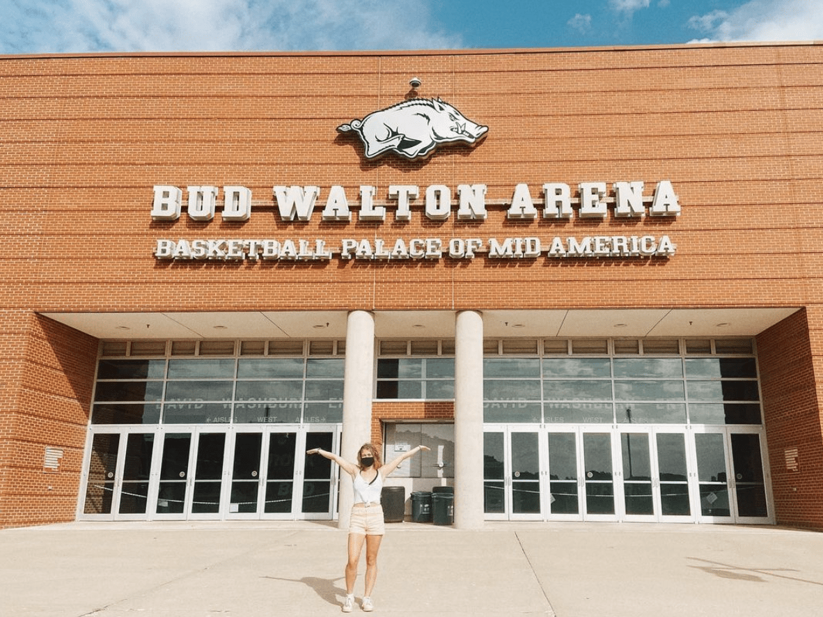 Billie Firmin poses in front of Bud Walton Arena, arms up to display the building, in 2020.